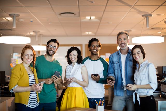 Diverse group of creative business professionals standing together in a modern office, all smiling and holding mobile phones. Ideal for use in articles or advertisements about teamwork, modern work environments, technology in the workplace, or diversity and inclusion in business settings.