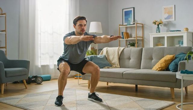 Fit man performing squat in bright living room home workout with dumbbells
