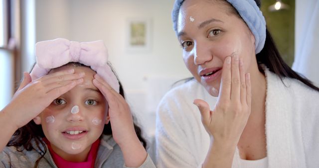 Mother and daughter applying face cream in bathroom. Both wear headbands and white gown while smiling and looking at camera. Perfect for promoting family bonding, skincare rituals, health, and beauty products.