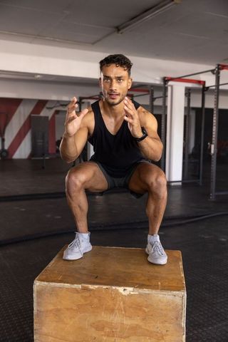 Young man performing low squat on plyo box in gym wearing smartwatch for strength training