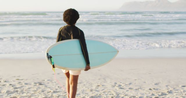 A young surfer holds a blue surfboard while standing on the beach at sunset. The ocean waves can be seen rolling onto the shore, with distant mountains visible. Perfect for illustrating themes of summer, outdoor recreation, surfing lifestyle, and youth adventure.