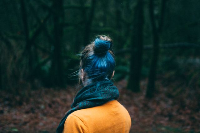 Woman with blue hair in a bun standing in a dusky forest. Wearing a scarf and autumn clothing. This can be used for concepts related to exploring nature, serenity, introspection, and unique fashion.