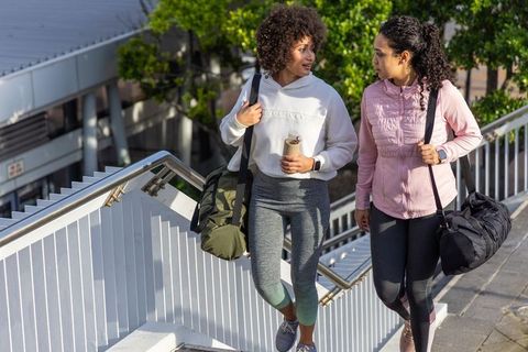 Two diverse women walking up stairs carrying gym bags and water bottle heading to gym