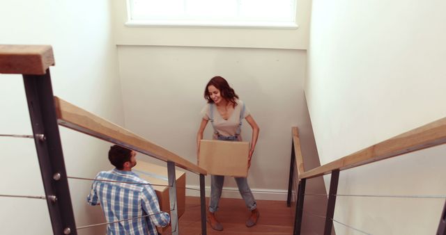 Couple moving cardboard boxes down staircase in their new home. Man handing box to woman as they relocate and unpack indoors. Ideal for themes related to moving homes, new beginnings, teamwork, and happiness in relocation.