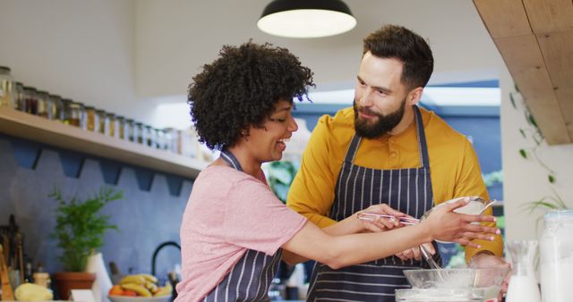 Image of happy diverse couple in aprons talking and baking together in kitchen at home. Happiness, inclusivity, free time, togetherness and domestic life.