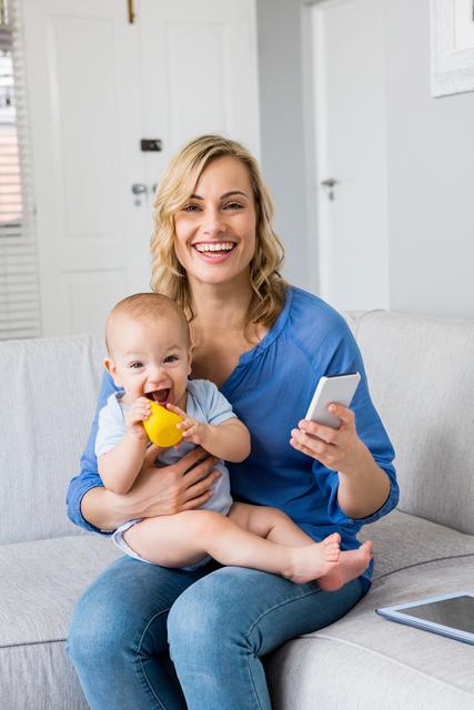 Smiling mother holding baby boy while using mobile phone in living room at home. Perfect for parenting blogs, family lifestyle articles, mobile technology advertisements, and home decor magazines.