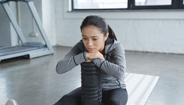 Focused asian woman using vertical foam roller on mat in bright minimalist gym studio