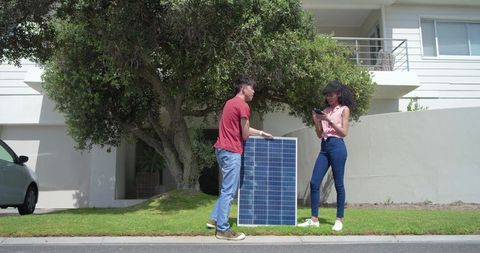 African american woman and asian man holding solar panel using smartphone on suburban lawn