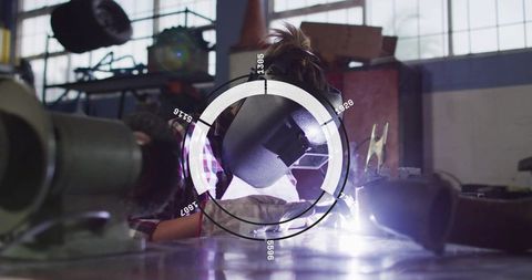 Female welder performing arc welding with helmet and hud overlay in gritty metal workshop