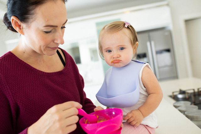 Caucasian mother feeding baby daughter making grumpy face sitting on counter top in kitchen. at home in isolation during quarantine lockdown.