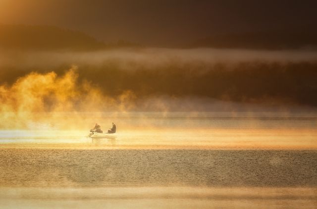 Two people enjoy fishing on a peaceful lake at sunrise, surrounded by mist and warm golden light. This serene scene is ideal for promoting outdoor activities, adventure tourism, and nature retreats. It captures the beauty of early morning tranquility and can be used in ads, brochures, and social media posts celebrating the great outdoors and relaxation.