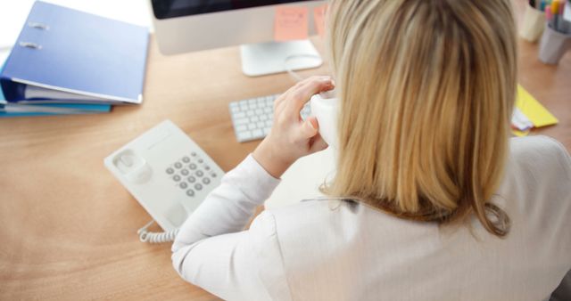 Young businesswoman with blonde hair using telephone while working at desk in a modern office setting. Ideal for business, corporate, or communication-themed promotions and presentations. Also suitable for usage in office-related articles, HR materials, and informational content about modern work environments.