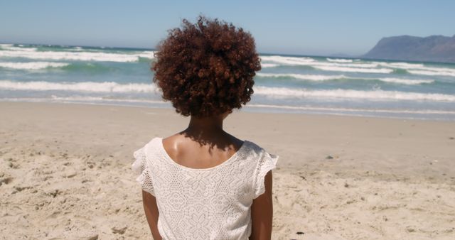 Woman with afro hairstyle standing on sandy beach, facing towards the ocean. Ideal for themes of relaxation, vacation, nature, solitude, self-reflection, and enjoying outdoor activities. Perfect for travel brochures, lifestyle blogs, and wellness articles.