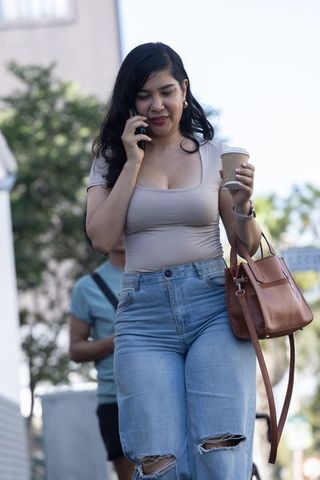 Young woman walking city sidewalk talking on phone holding coffee and brown handbag in ripped denim