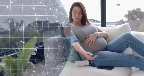 Pregnant woman monitoring blood pressure on sofa with digital cuff and health network overlay