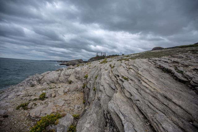 Stunning coastal landscape featuring rocky terrain under a dramatic, cloudy sky. Ocean waves crash against rocks, while distant greenery contrasts with rugged stone. Perfect for use in travel brochures, nature photography portfolios, geology studies, or inspiring adventure-themed media.