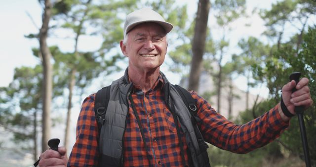 Senior man enjoying a hike in a forest while smiling and holding trekking poles. Ideal for use in articles about active aging, outdoor activities for seniors, healthy lifestyles, and adventure.