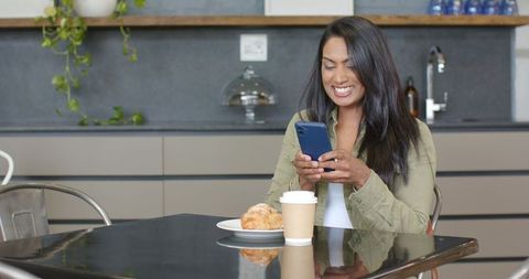 Mid adult indian woman smiling while texting on phone with coffee and croissant in modern kitchen