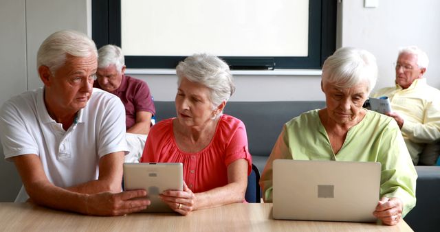 Elderly individuals sitting together while using tablets and laptops, focusing on technological gadgets. Ideal for content related to senior education, digital literacy programs, and promoting technology use among older adults.