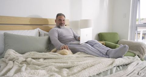 Senior Man Relaxing on Bed with Laptop and Petting Dog at Home