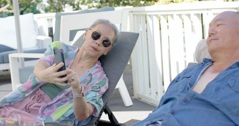 Asian senior couple relaxing on sunlit deck loungers woman using smartphone, man resting