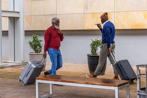 Two travelers walking with rolling luggage in urban plaza, using phone and headphones