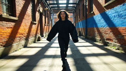 Woman striding through sunlit urban alleyway with dramatic shadow bands across brick corridor