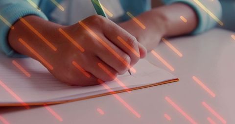 Closeup hand writing in orange-bound notebook with green pencil and warm light streaks