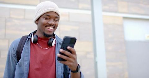 Smiling African American Man Checking Smartphone Outdoors with Beanie Headphones Backpack