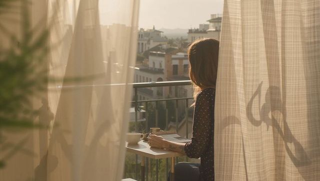 Woman on sunlit balcony sipping coffee with newspaper at golden hour, urban calm