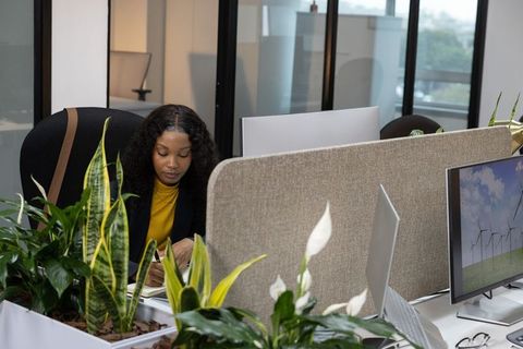 African American woman writing at modern partition desk surrounded by plants and monitors
