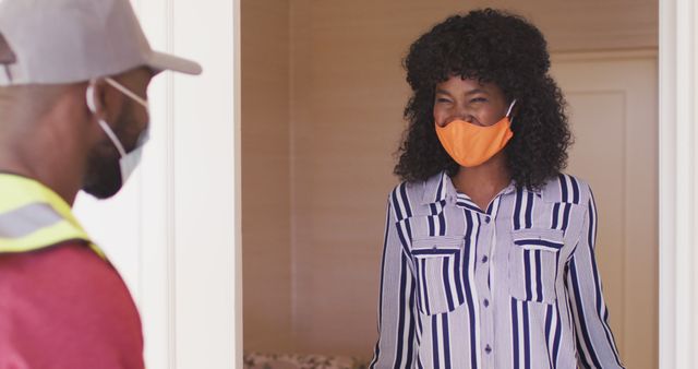 Woman standing at her doorway wearing an orange face mask and stripe shirt, receiving a delivery from a person in a work uniform also wearing a mask. Suitable for use in topics related to pandemic safety, delivery services, contactless interactions, home services, and customer service.