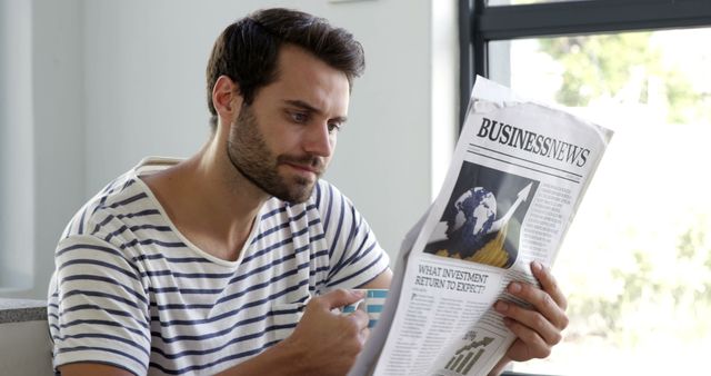 Young man reading a business section of a newspaper in a relaxed indoor environment. Perfect for use in topics related to finance, morning routines, casual living, and business-oriented content.