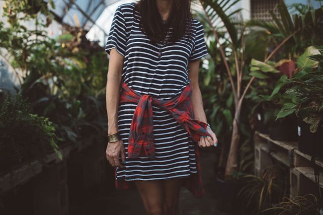 Woman wearing striped dress with plaid shirt wrapped around waist standing in greenhouse filled with various plants. Wrist jewelry adds a casual, bohemian touch. Perfect for use in fashion, lifestyle, gardening, and environmental themes.