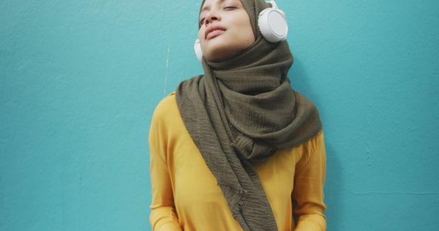 Young Muslim woman in hijab wearing white headphones and yellow blouse, enjoying music against teal background. Perfect for use in lifestyle, fashion, technology, and modern culture contexts. Highlights themes of relaxation, trendy casual wear, and multicultural expression.