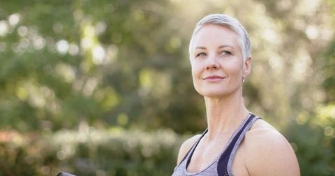 Confident middle-aged woman gazing outdoors holding yoga mat in park promoting wellness