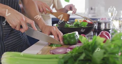 Couple cooking together chopping green pepper and stirring leafy greens on home stovetop