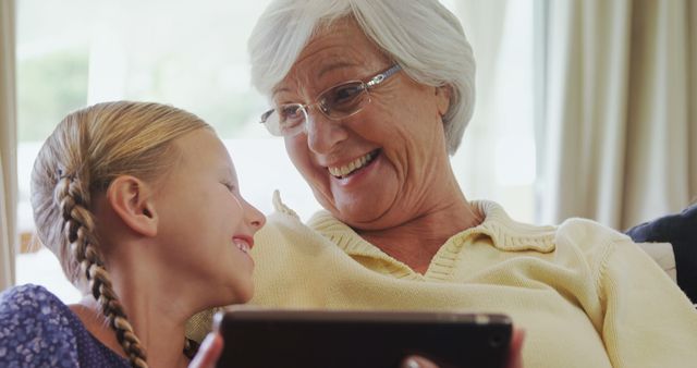 A senior Caucasian woman and a young girl share a joyful moment looking at a tablet, with copy space. Their smiles suggest a close bond, a grandmother enjoying technology with her granddaughter.