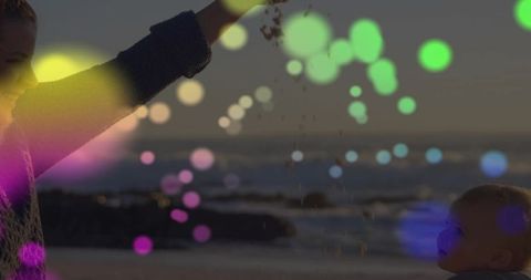 Caregiver lifting arm releasing sand at dusk beach with colorful bokeh light effects