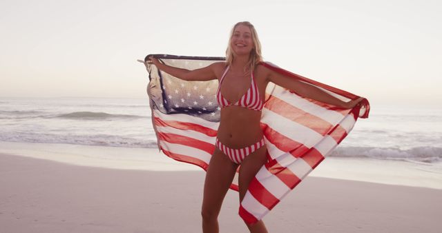 Portrait of happy caucasian woman holding flag of usa and smiling on beach. Patriotism, lifestyle, realxation, free time and vacation.