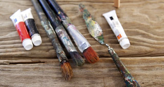 This depicts several worn paintbrushes and paint tubes scattered on a rustic wooden surface. It highlights the tools used by artists. Suitable for content related to art, creative processes, DIY projects, and artistic supplies.
