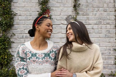 Two women wearing festive headbands sharing warm laughter in cozy winter sweaters