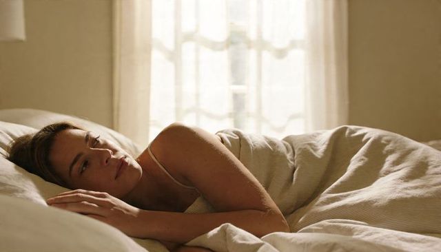 Serene morning woman resting in bed with soft daylight, neutral linens and calm expression