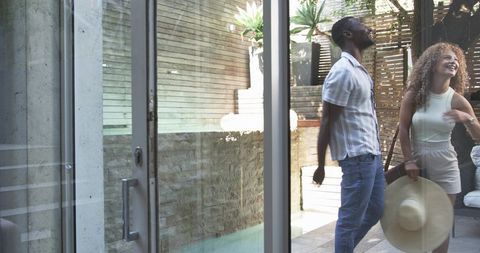 Sunlit courtyard couple laughing through sliding glass door holding straw sunhat, modern