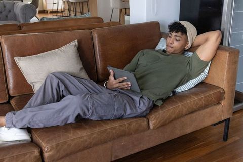 African american man relaxing on brown leather sofa holding tablet in modern living room