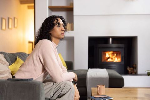 Non-binary person sitting on gray sofa gazing at fireplace in cozy modern living room