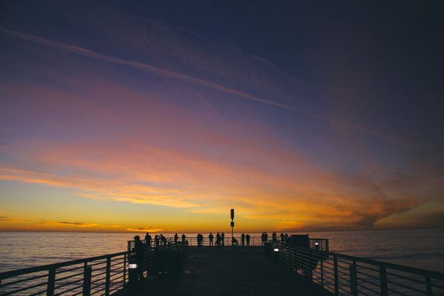 A group of people are standing on a pier overlooking the ocean as the sun sets, creating a beautiful array of colors in the sky. The calm water and twilight ambiance give a serene and tranquil feeling. This image is perfect for travel promotions, serene nature backdrops, and tourism advertisements suggesting peaceful and picturesque destinations.
