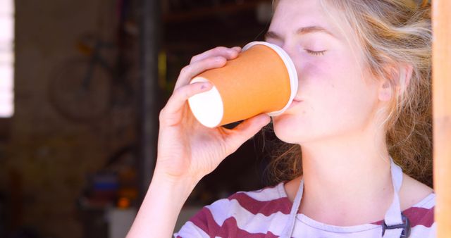 A young woman enjoying a hot beverage from a paper cup in a casual home setting. She wears a striped shirt and looks content and relaxed. Image captures a moment of personal enjoyment and comfort, ideal for use in advertisements for beverages, lifestyle blogs, home living articles, and relaxation or wellness content.