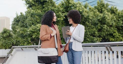 Diverse female colleagues sharing tablet on urban bridge, casual business conversation