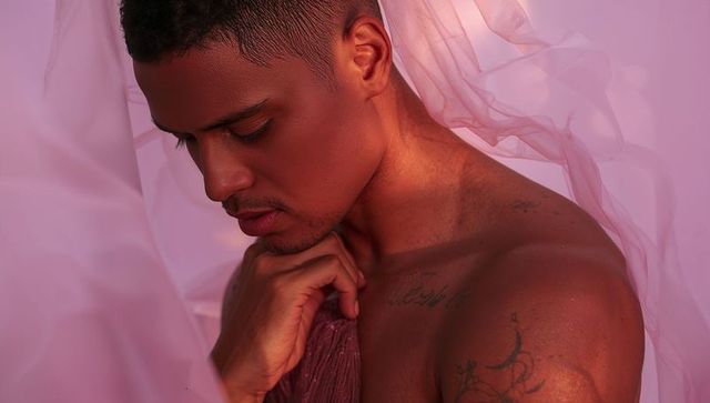 Intimate shirtless tattooed man clutching sheer fabric under soft pink moody studio light